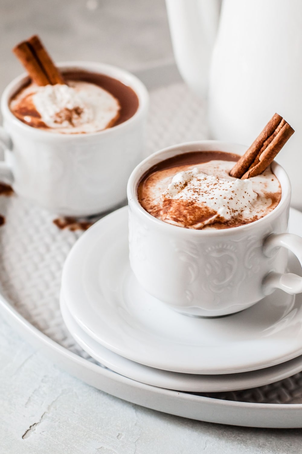 Two servings of Mexican hot chocolate on a white mug with cinnamon sticks and cream. 