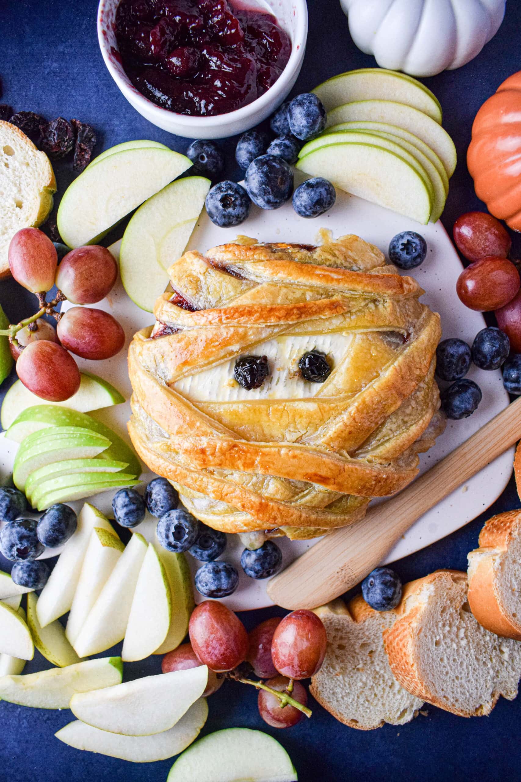 Puff pastry with a creamy Brie filling, adorned with two berry eyes, surrounded by apple slices and bread on a white plate.