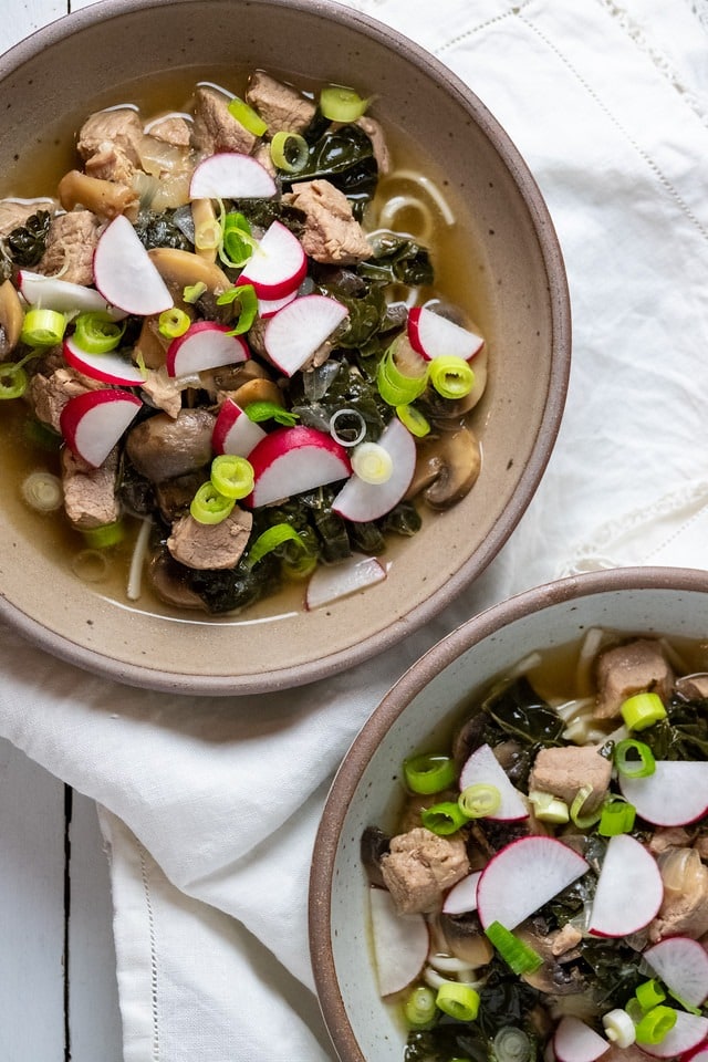 Two bowls of soup with pork and vegetables, served hot and garnished with fresh herbs
