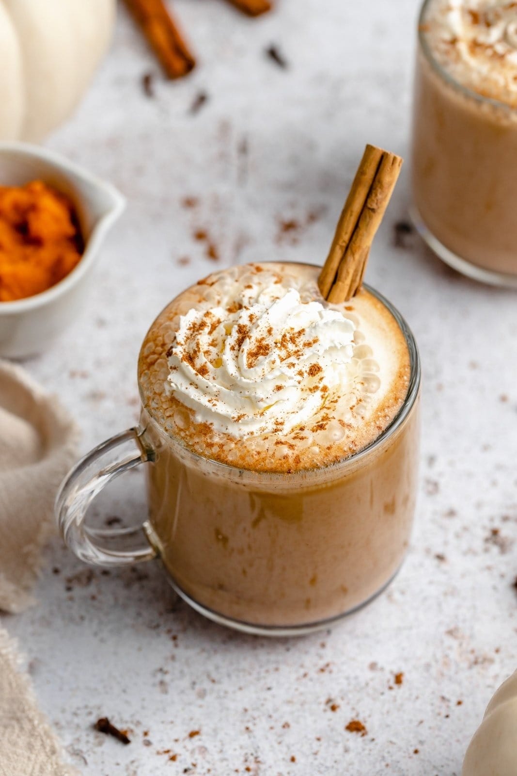 Glass mug of homemade pumpkin spice latte topped with cream frosting cinnamon sticks and powder. 