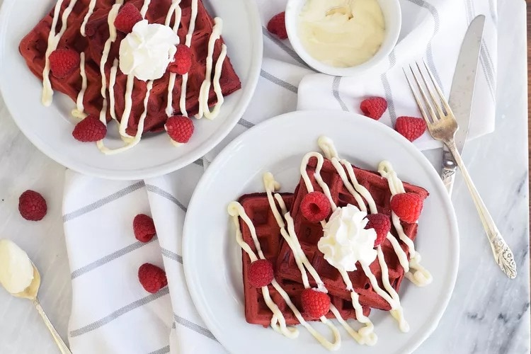 Top view of two plate with red velvet flavored waffles with raspberries drizzled with white chocolate and whipped cream on top,