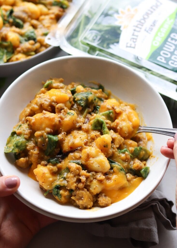 Hand holding a bowl of sausage and gnocchi with creamy butternut squash sauce and baby greens. 