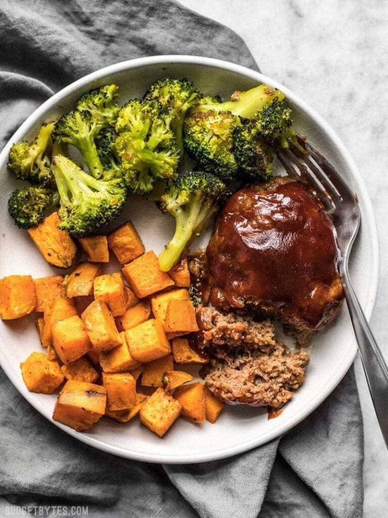 Sliced BBQ meatloaf topped with gravy served on a plate with broccoli, and roasted potatoes. 