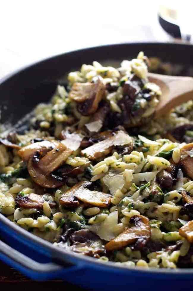 Creamy mushroom and spinach orzo on a blue  pan tossed by a wooden spoon. 