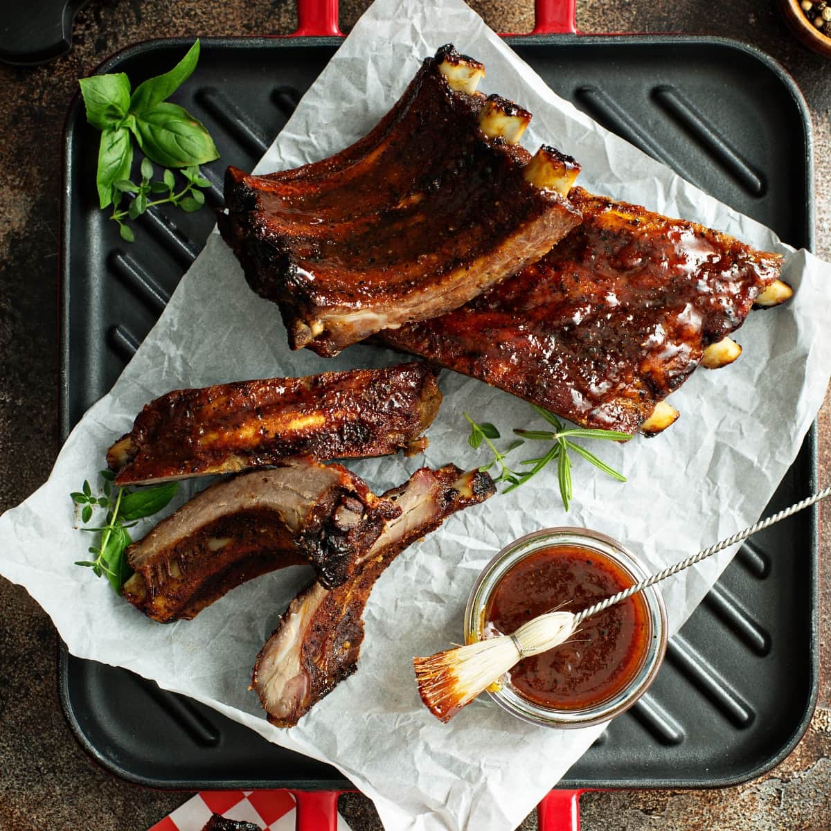 Different Types of Ribs Explained featuring Top View of Cooked, Bone-In Ribs with Barbecue Sauce on Parchment and a Cast Iron Grill