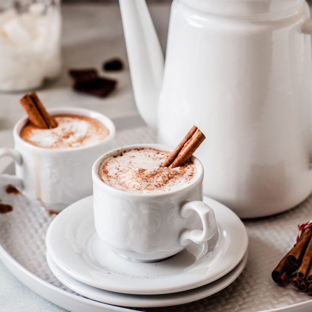 Two servings of Mexican hot chocolate in a white mug and pitcher garnished with cinnamon sticks and cream. 