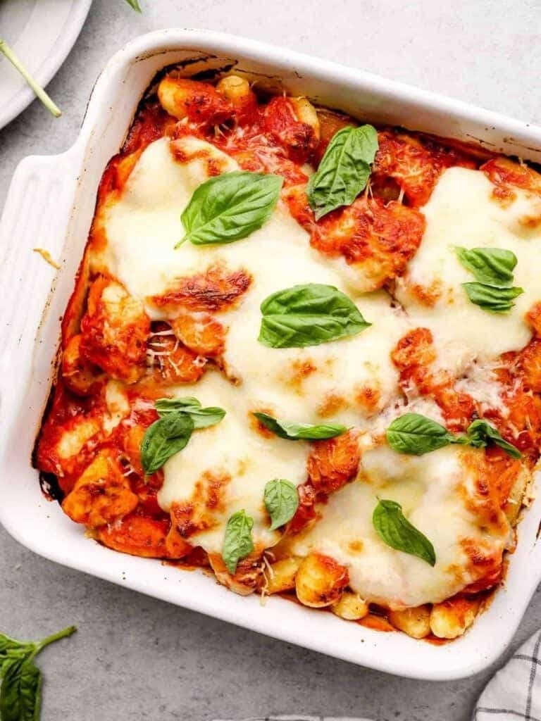 Chicken parmesan gnocchi bake on a white baking dish garnished with fresh spinach leaves. 