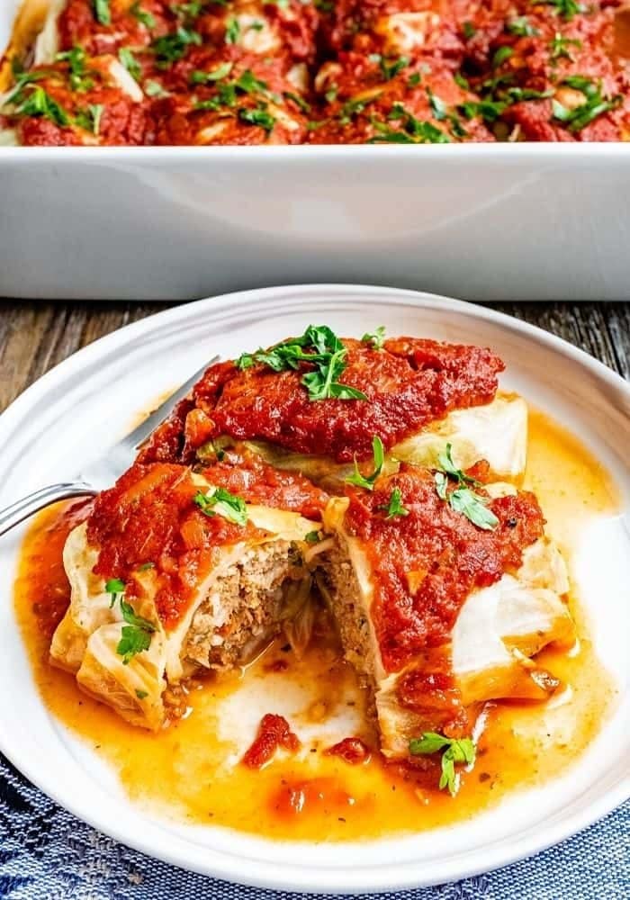 Ground beef cabbage rolls topped with tomato sauce and herbs served on a white plate with fork. 
