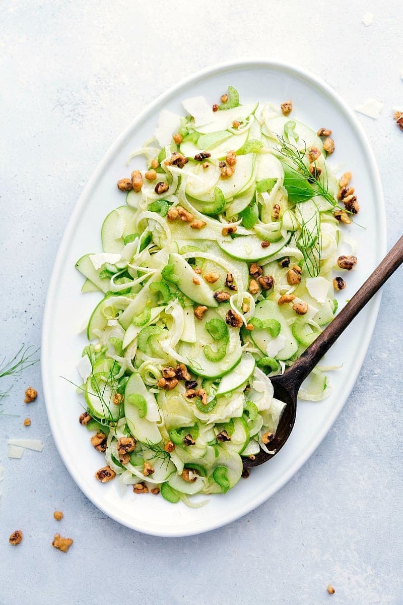 A Plate of Apple Fennel Salad with thinly sliced apples, celery, and fennel topped with walnuts and parmesan served with a wooden spoon