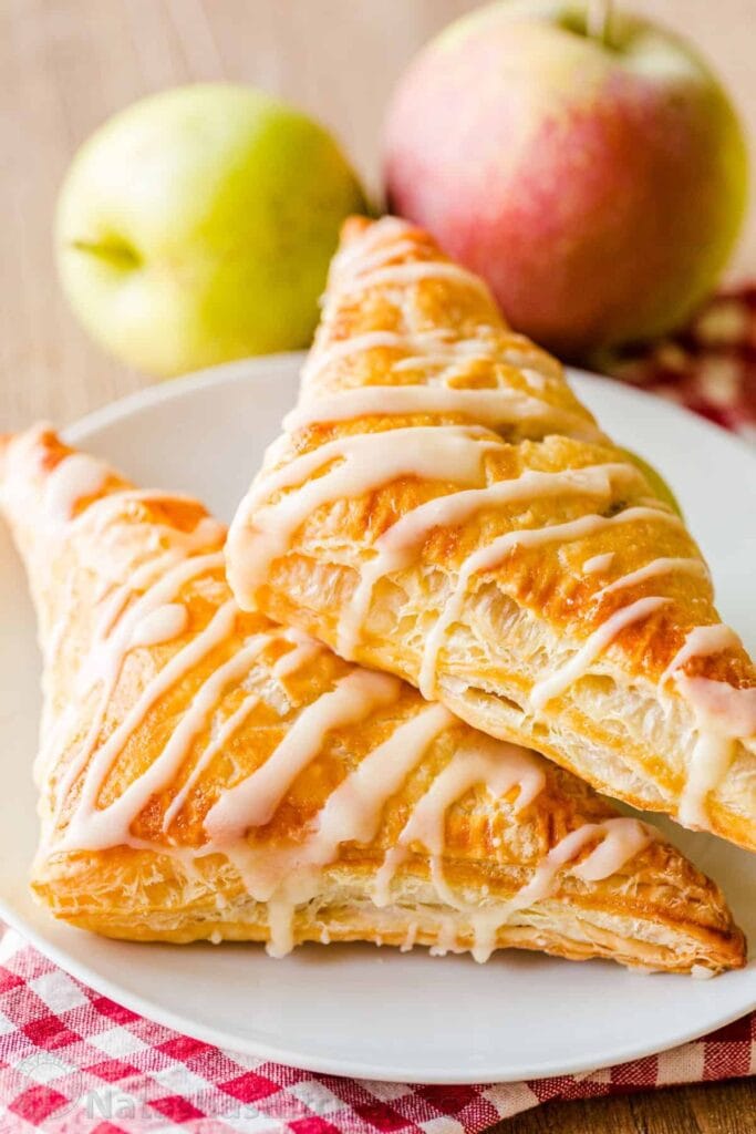 Apple turnovers on a plate drizzled with sugar glaze and whole apples in the background. 