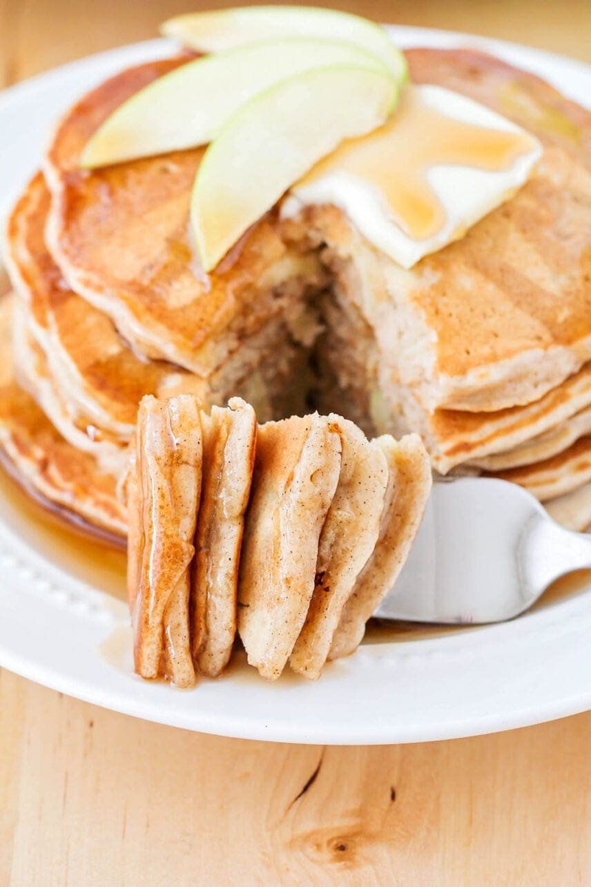 Stack of pancakes topped with butter, maple syrup and and apple slices with a slice pierced with a fork.