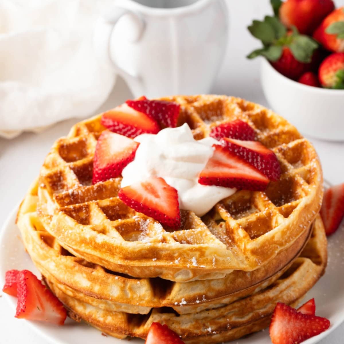 Stack of Belgian Waffles with powdered sugar, whipped cream and sliced strawberries on top, white bowl with strawberries in the background