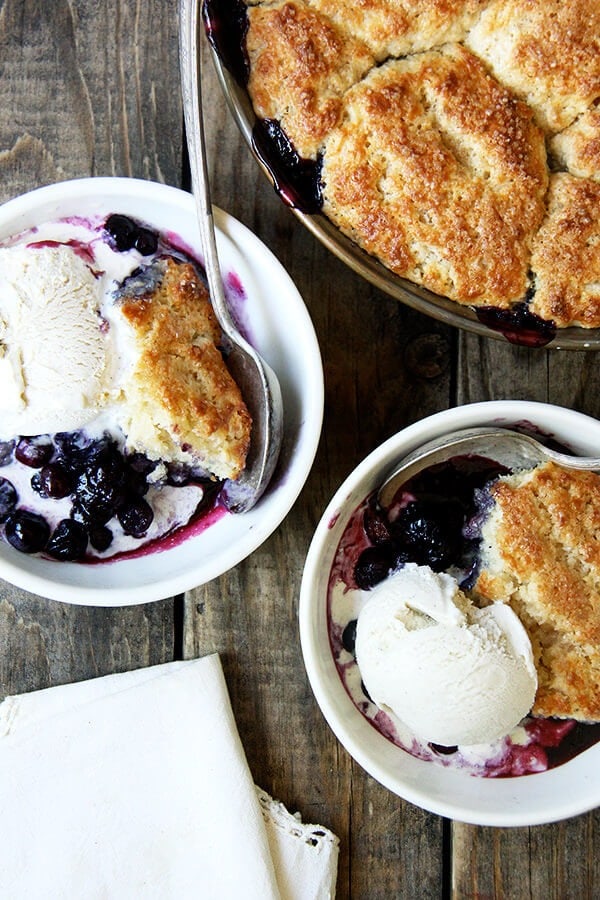 Two bowls of Blueberry Cobbler with ice cream top and a pan of freshly baked Blueberry Cobbler
