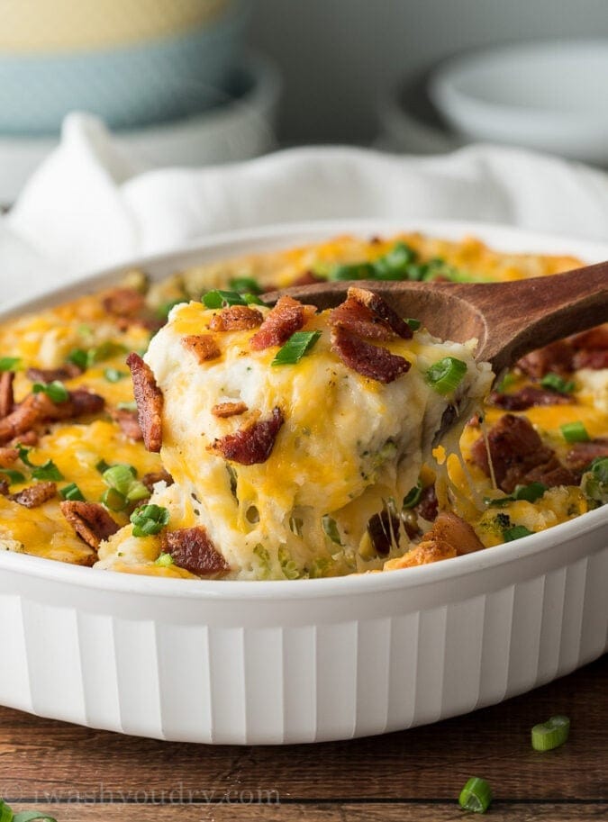 Wooden spoon scooping a portion of a gooey mashed potato casserole with bacon bits and chopped onion leaves. 
