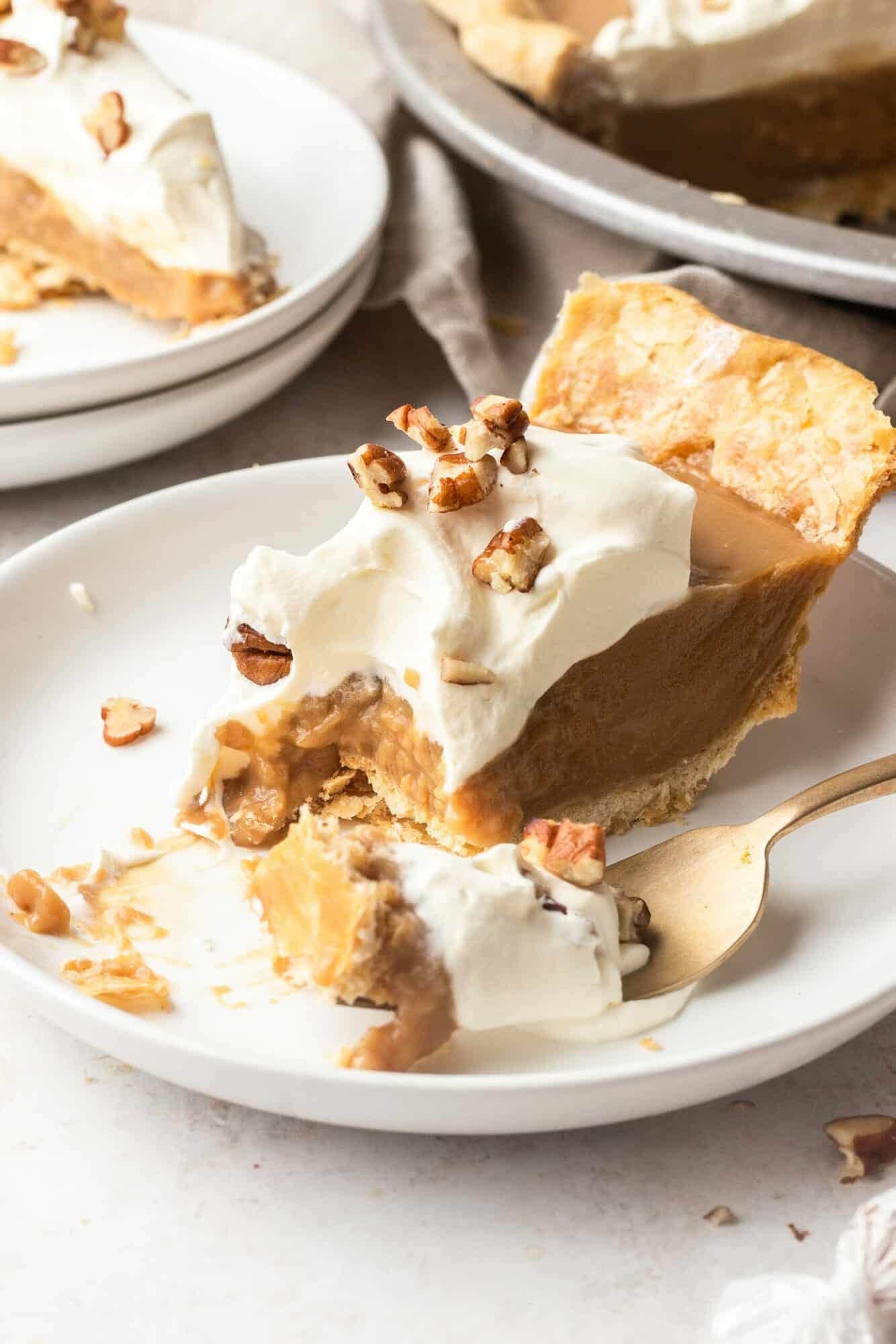 A slice of caramel pie with thick filling and whipped cream and chopped nut on top sliced with a fork. 