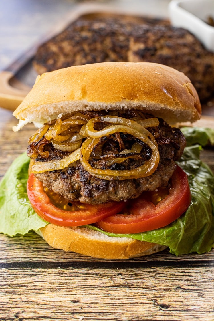 Burger with thick patty, tomato, lettuce and caramelized onions filling on top of a wooden table.  