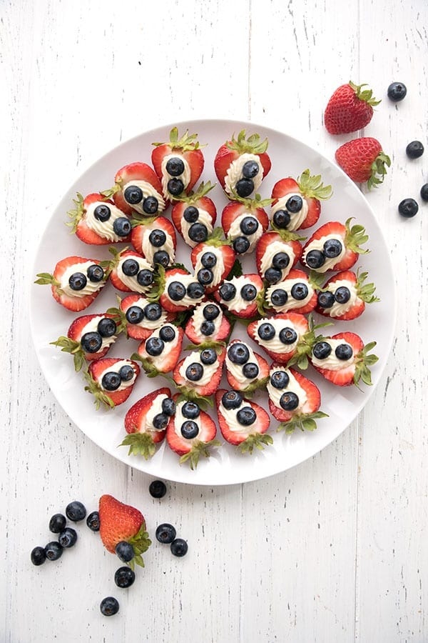 Strawberries sliced in half arranged in a white plate decorated with cheesecake cream and berries. 