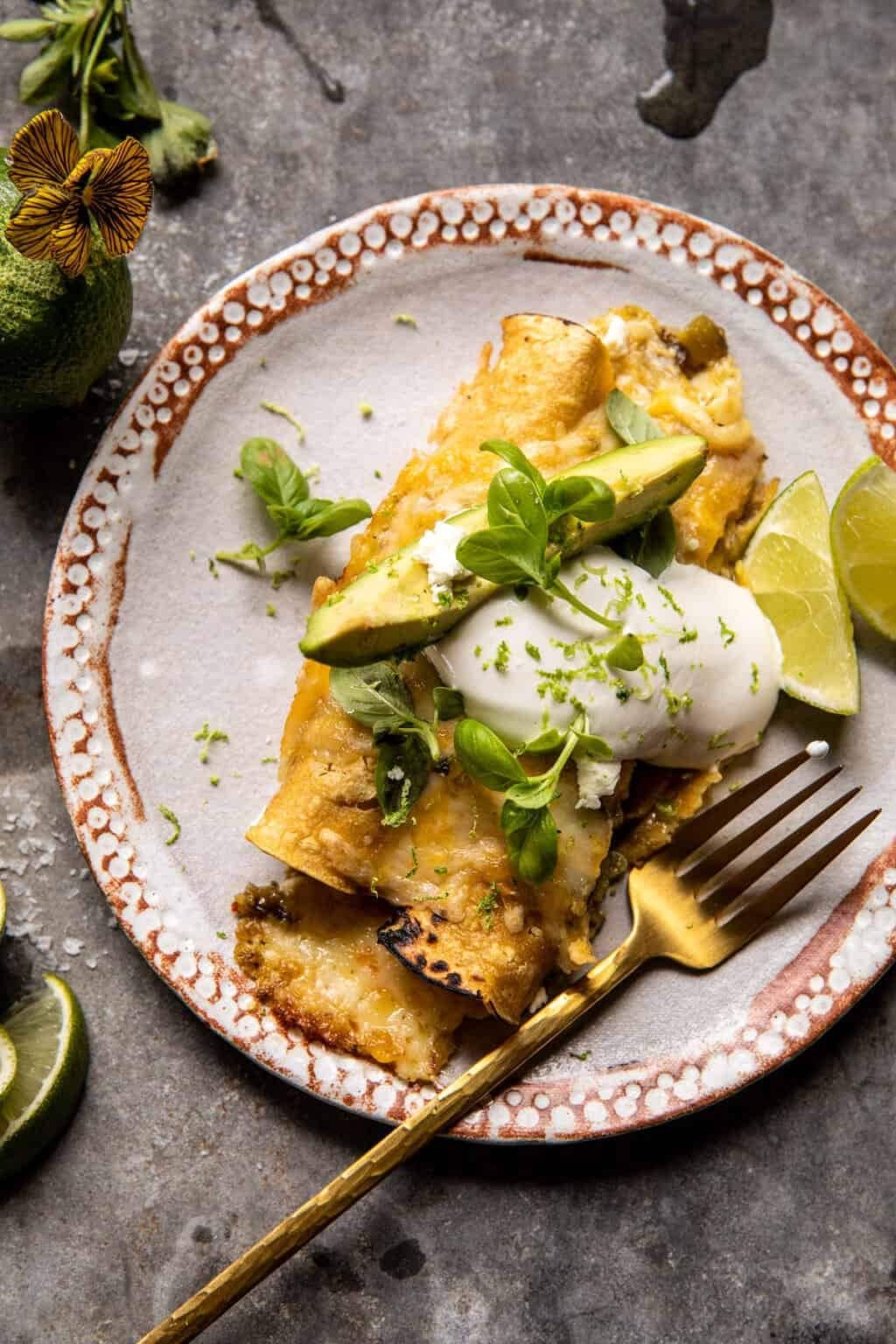 Corn enchiladas topped with sour cream garnished with avocado and lemon slice and fresh herbs.