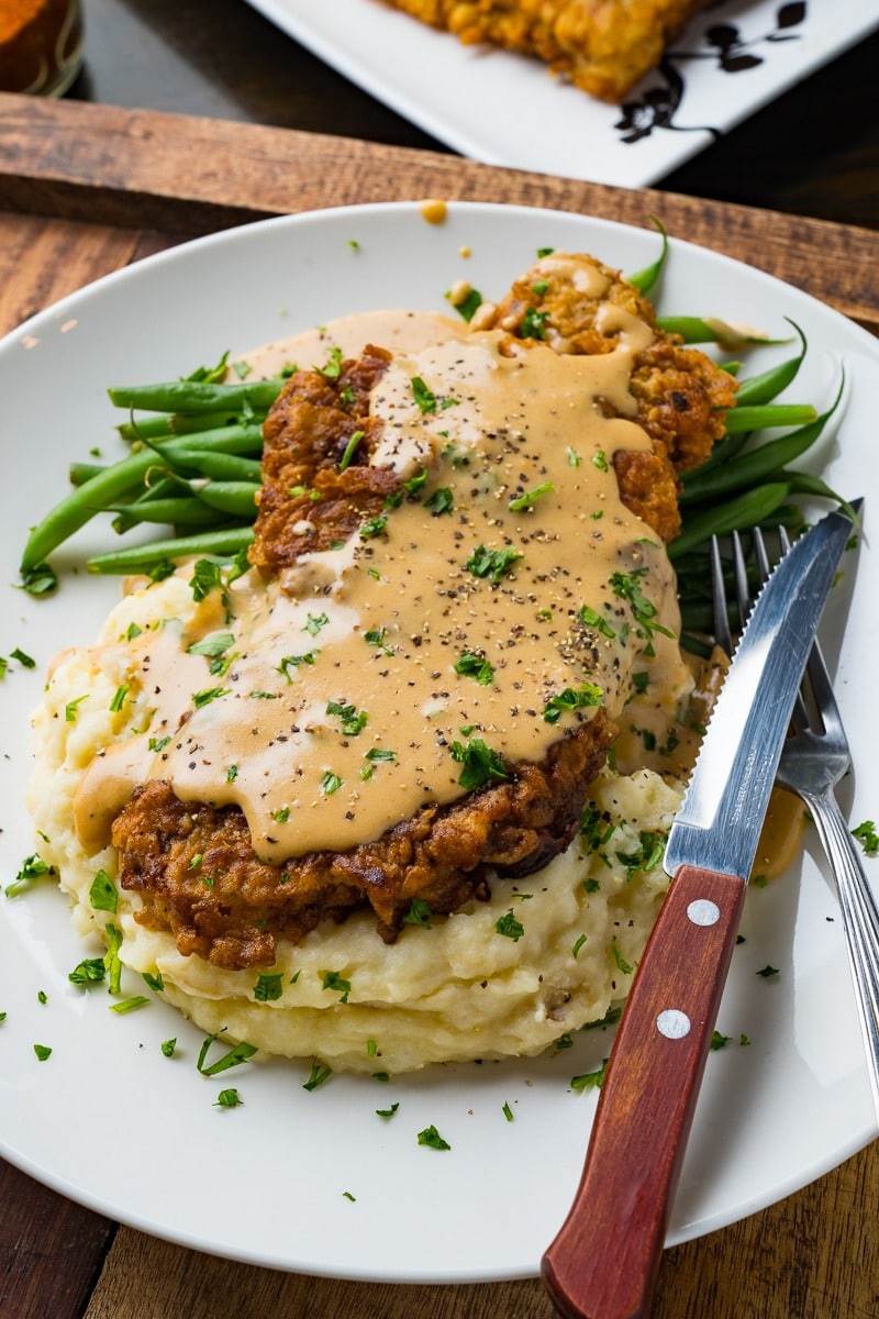 Fried steak on top of mashed potatoes and boiled green beans served on a plate with gravy on top