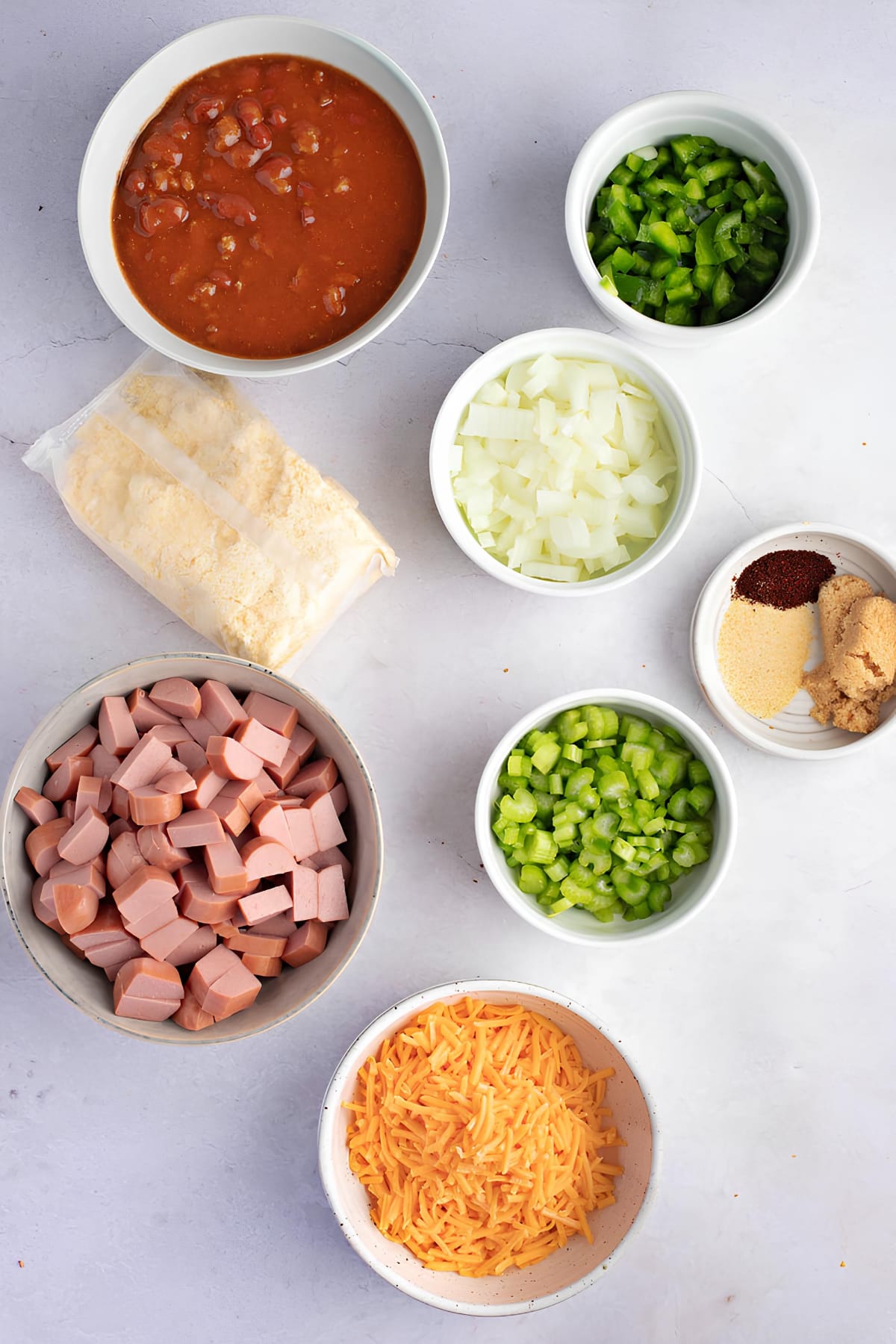 Bowls of chopped green pepper, onion, celery, chili with beans, bite-sized hotdogs, shredded cheese, a saucer of garlic powder, chili powder, sugar and a pack of cornbread or muffin mix
