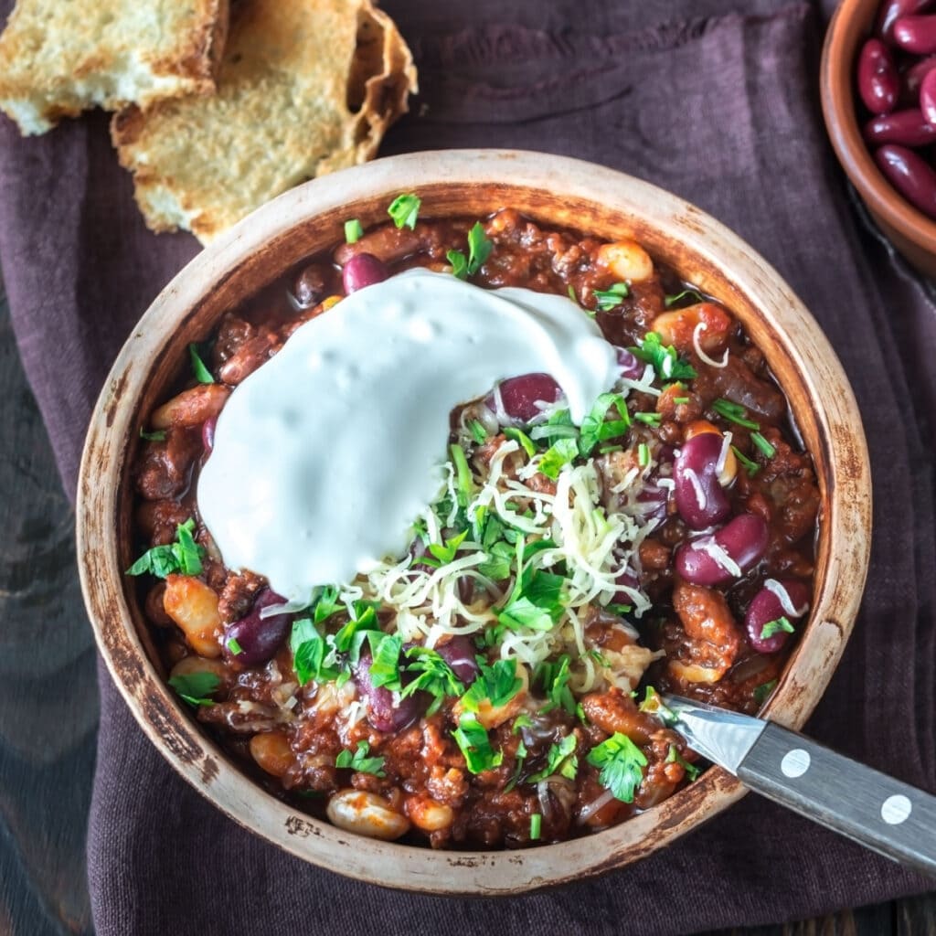 Chili in a wooden bowl with cheese, sour cream and chopped parsley toppings. 