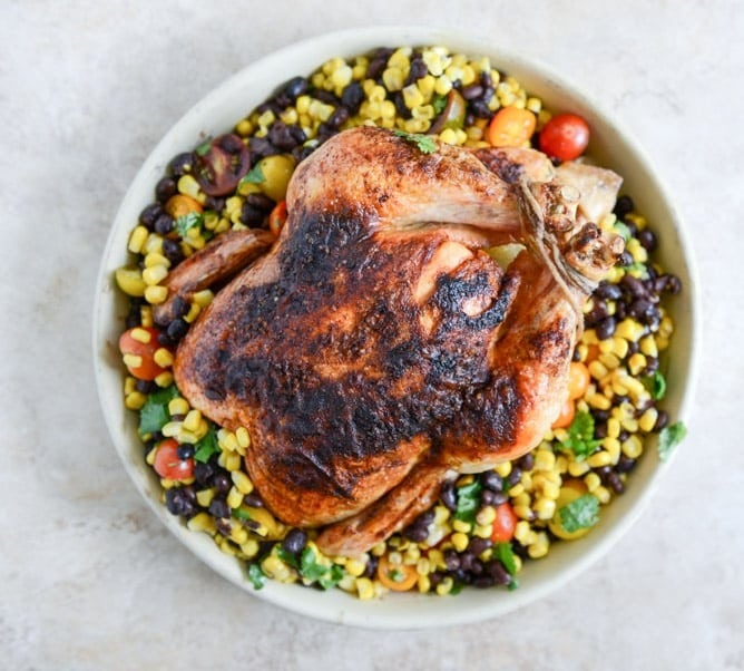 Whole roasted chicken rested on top of a black bean corn salad on a bowl. 