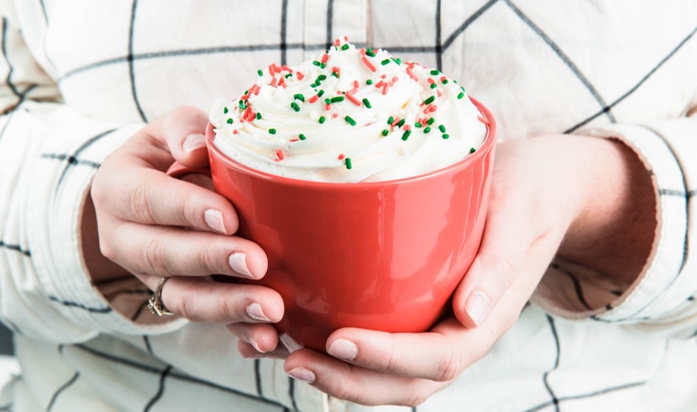 Hand holding a red mug filled with coffee topped with whipped cream and garnished with sprinkles.
