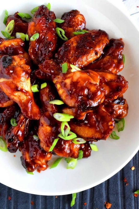 Top view of a chicken wings coated with sticky sauce garnished with chopped chili served on a white plate.