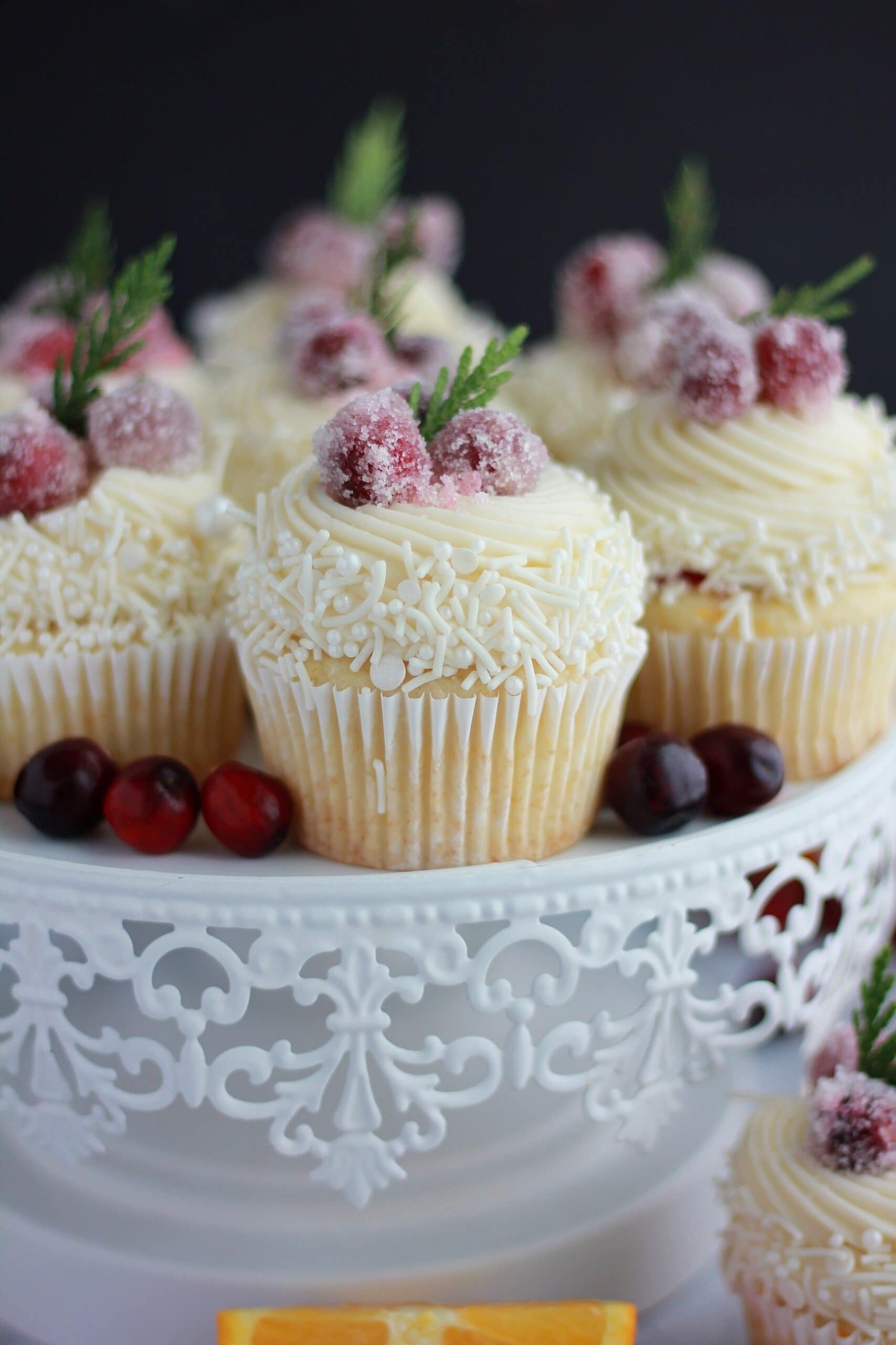 White chocolate cupcakes topped with frosting, cranberry compote and sugared cranberries on a cake tray. 