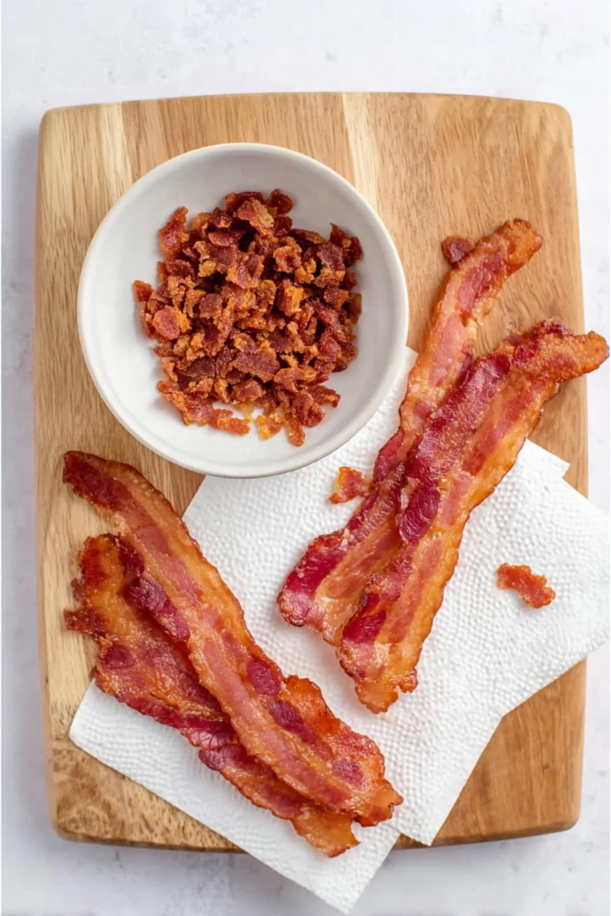 Crispy cooked bacon strips on paper towels with a small bowl of crumbled bacon on a wooden cutting board.