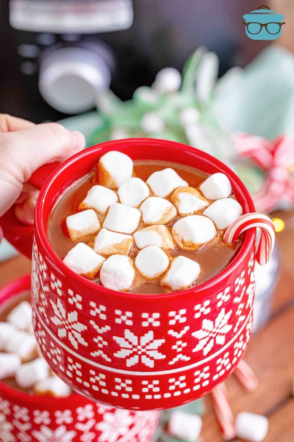 Hand holding a holiday mug with hot chocolate topped with marshmallows and candy cane.