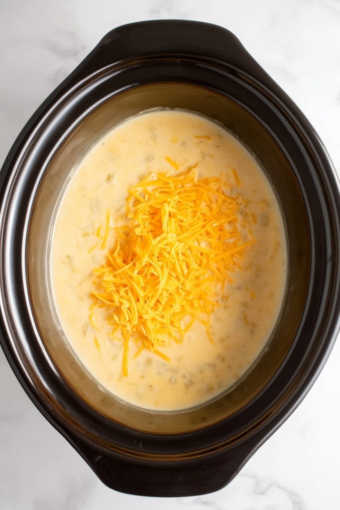 A slow cooker filled with creamy potato soup topped with a pile of shredded cheddar cheese.