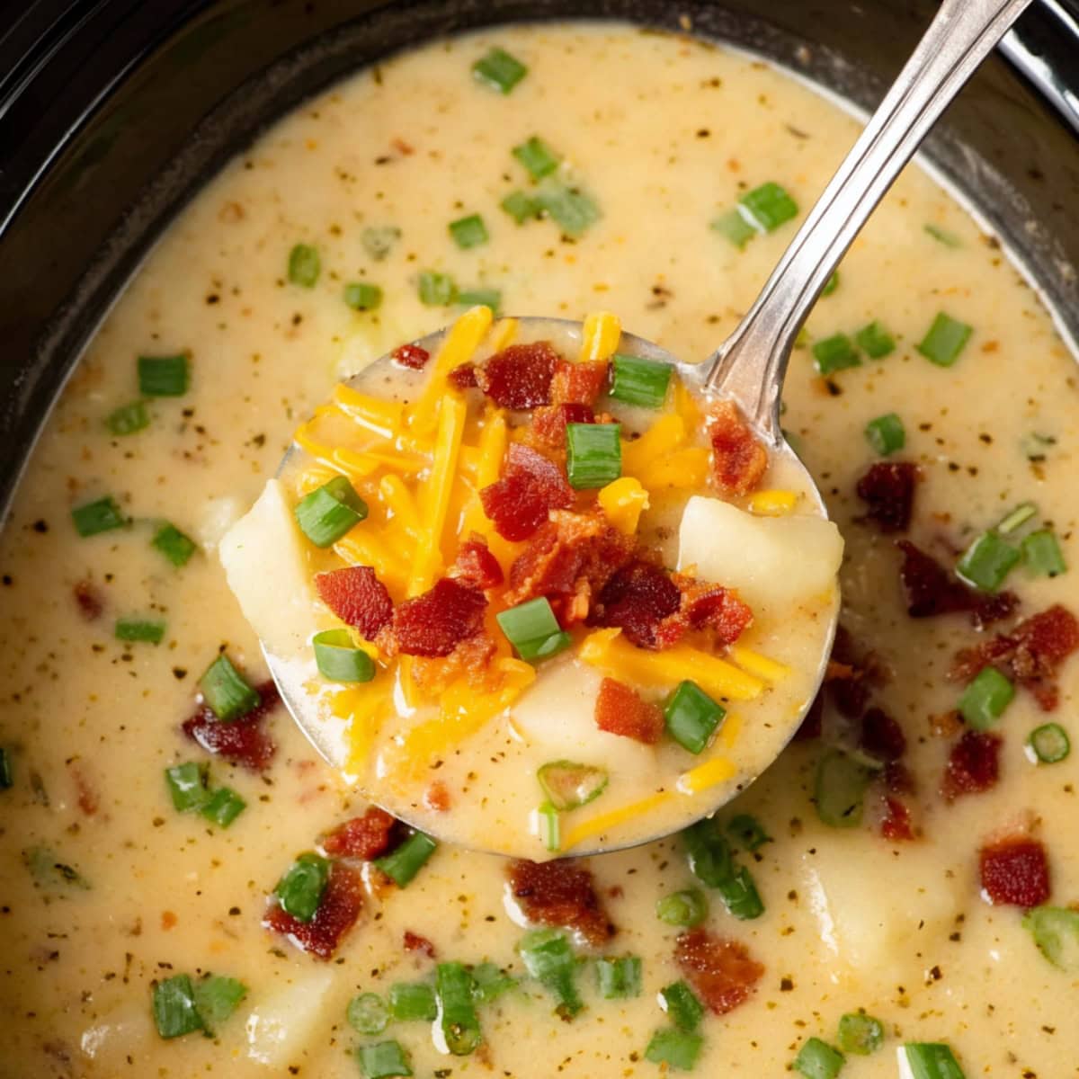 Close-up of a ladle of potato soup with bacon, cheese and green onions in a black slow cooker.