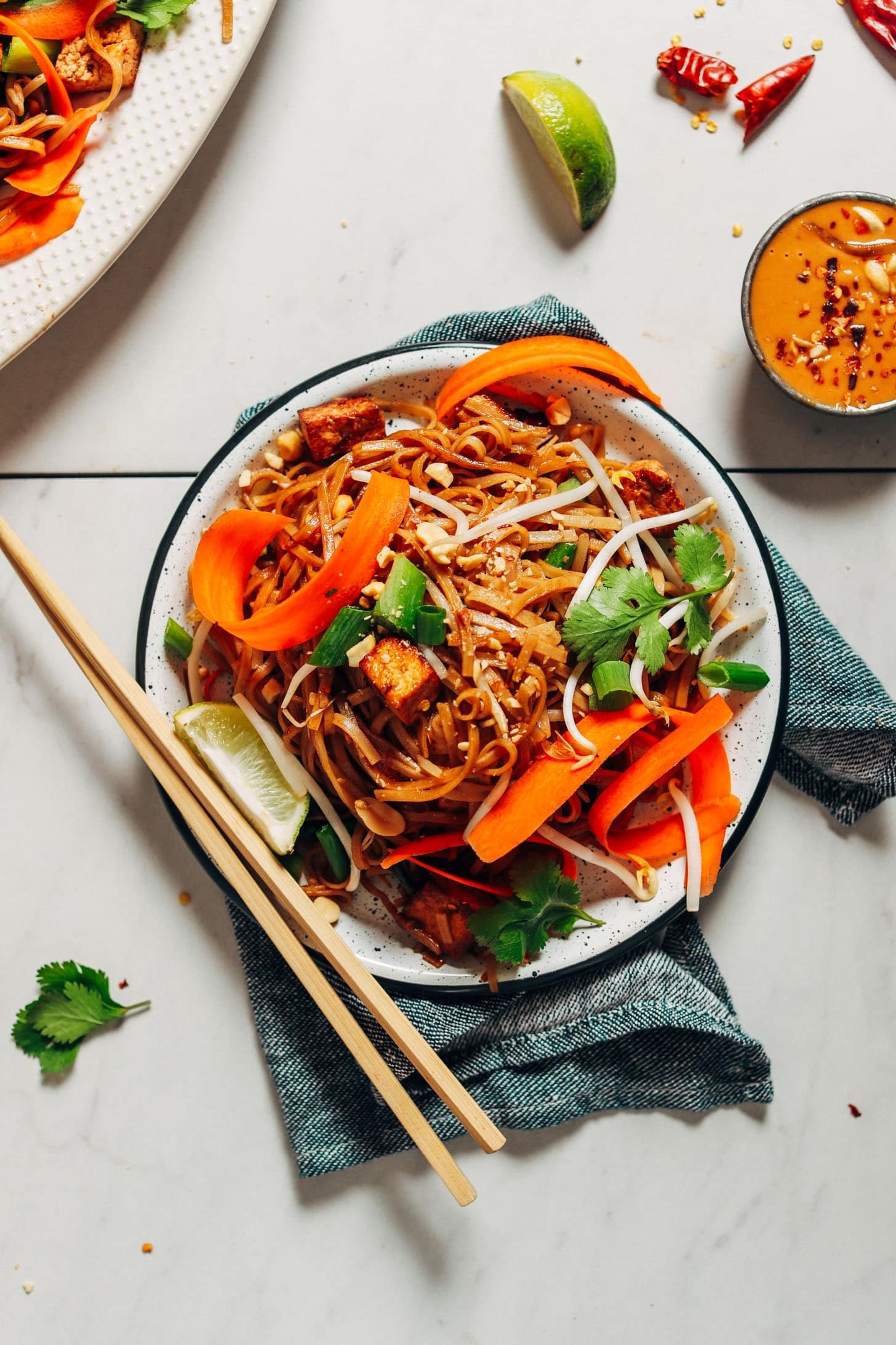 A serving of pad Thai on a plate garnished with lime wedges, bean sprouts, peanut sauce, shredded carrot, cilantro, and sriracha or chili garlic sauce.