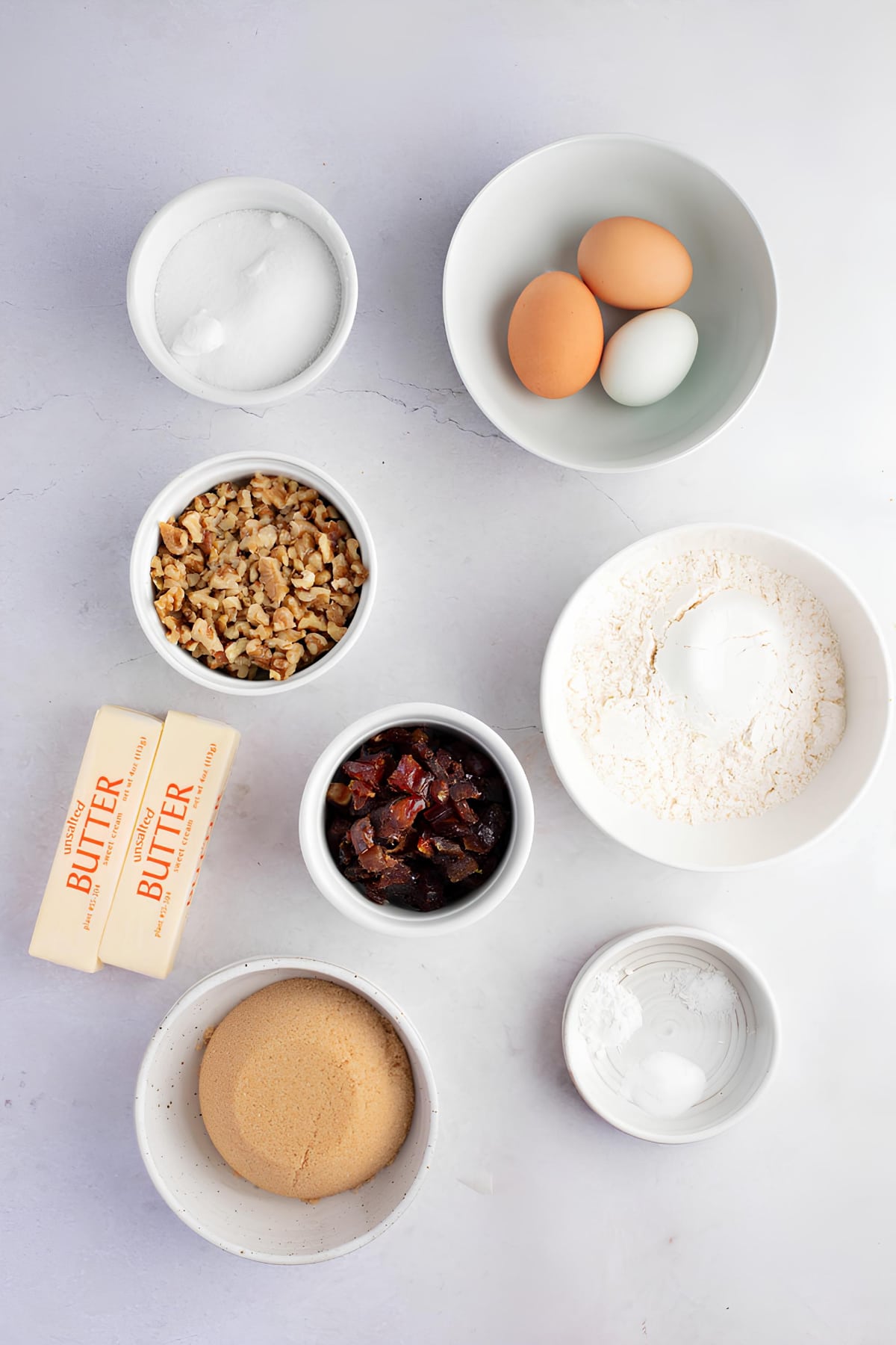 Food for the Gods ingredients- eggs, sugar, dates, chopped walnuts, butter and flour flat lay on a white table surface.