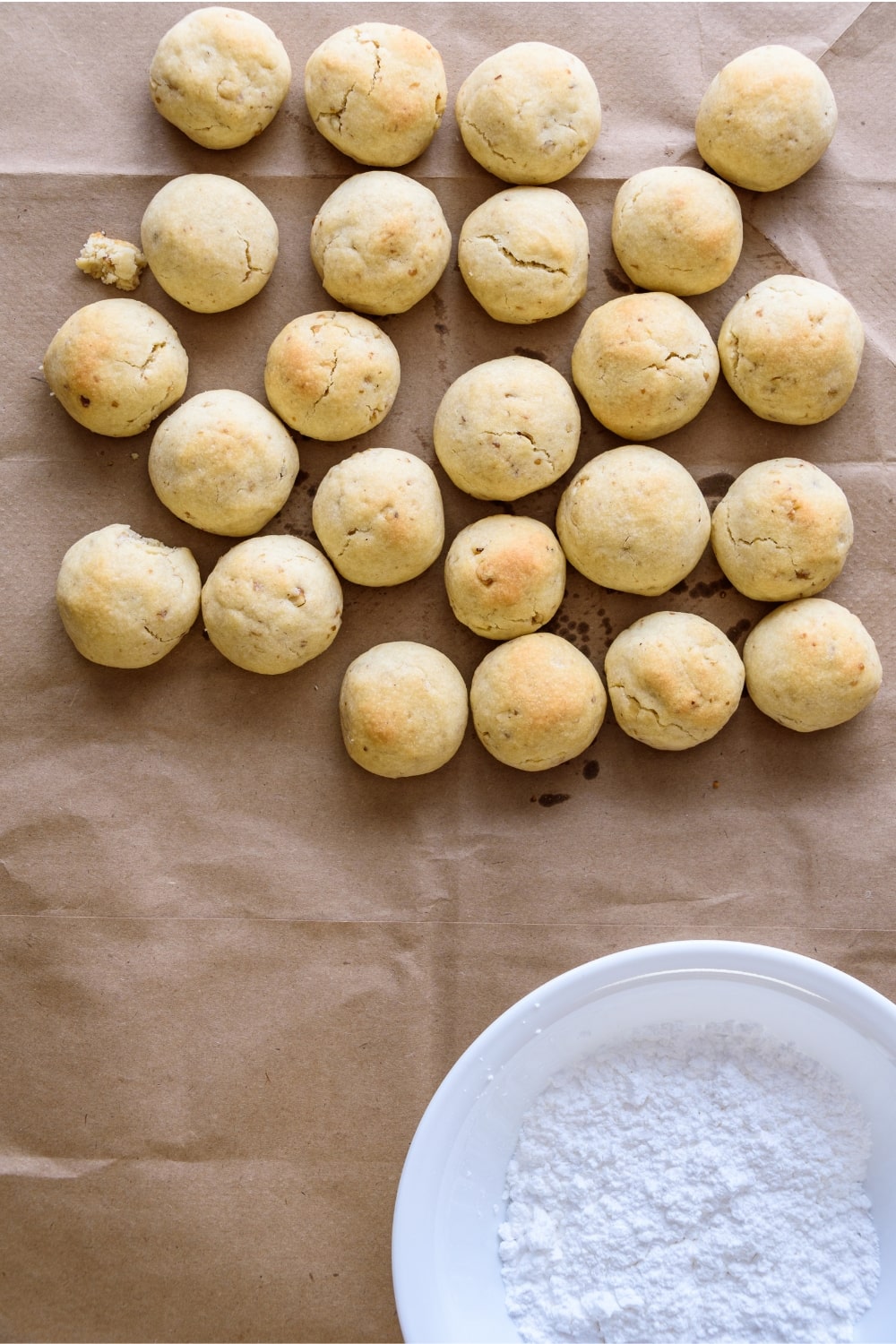 Fresh baked Italian wedding cookies on parchment paper and bowl of powdered sugar ready to be coated.