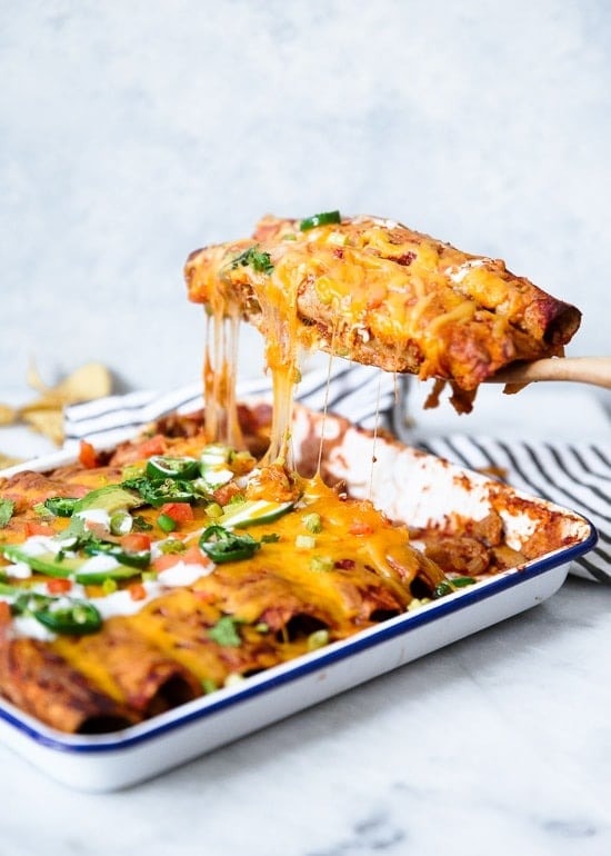 Wooden spoon scooping a portion of cheesy chicken enchilada in a baking pan. 