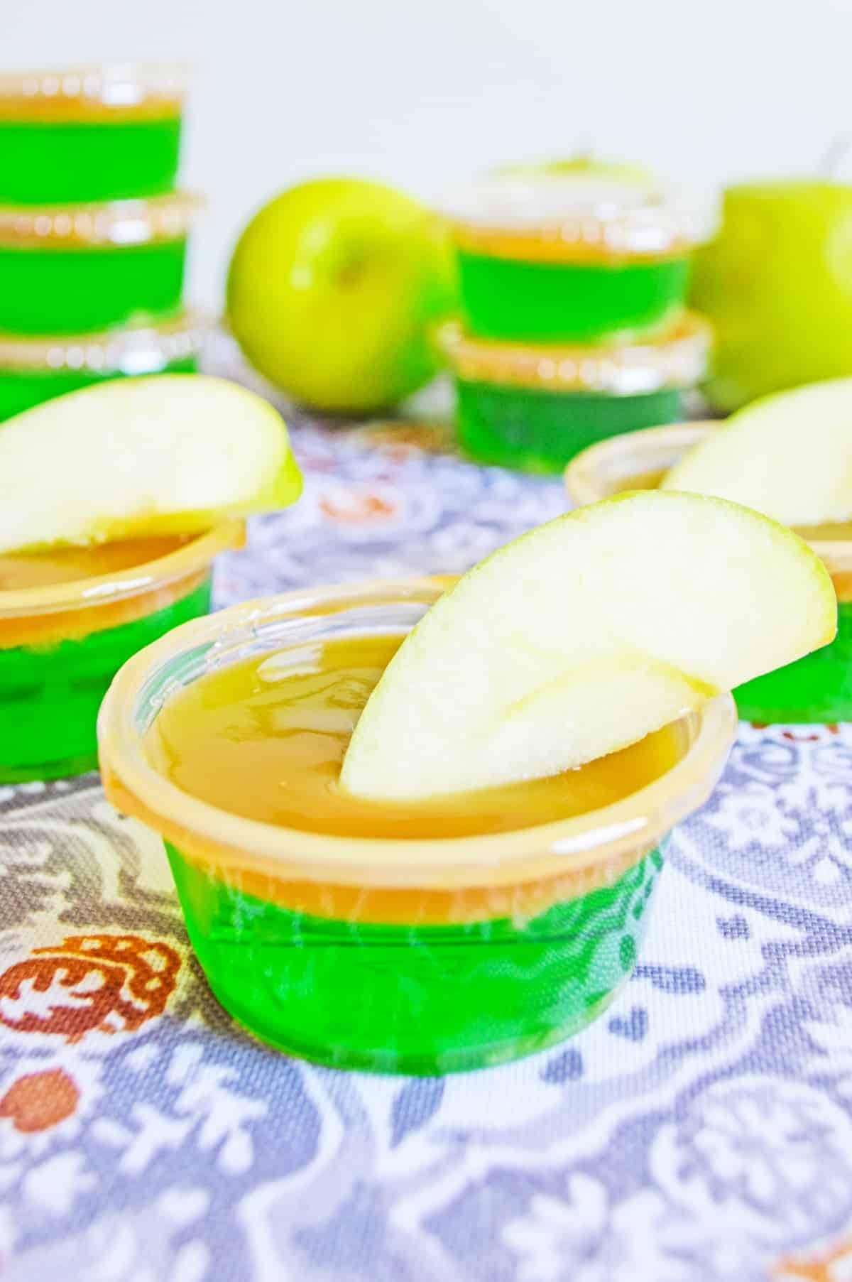 Apple Jello shots on plastic cups topped with caramel syrup and apple slice garnish.