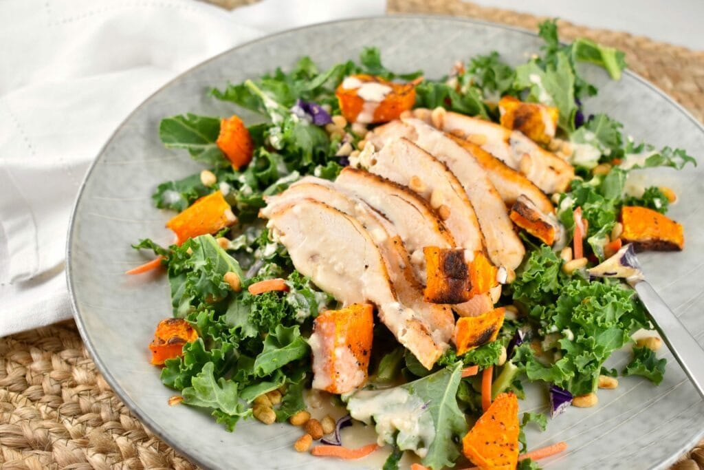 A plate of fresh kale greens, topped with grilled chicken, roasted butternut squash, drizzled with a zesty lemon tahini dressing, and topped with toasted pine nuts
