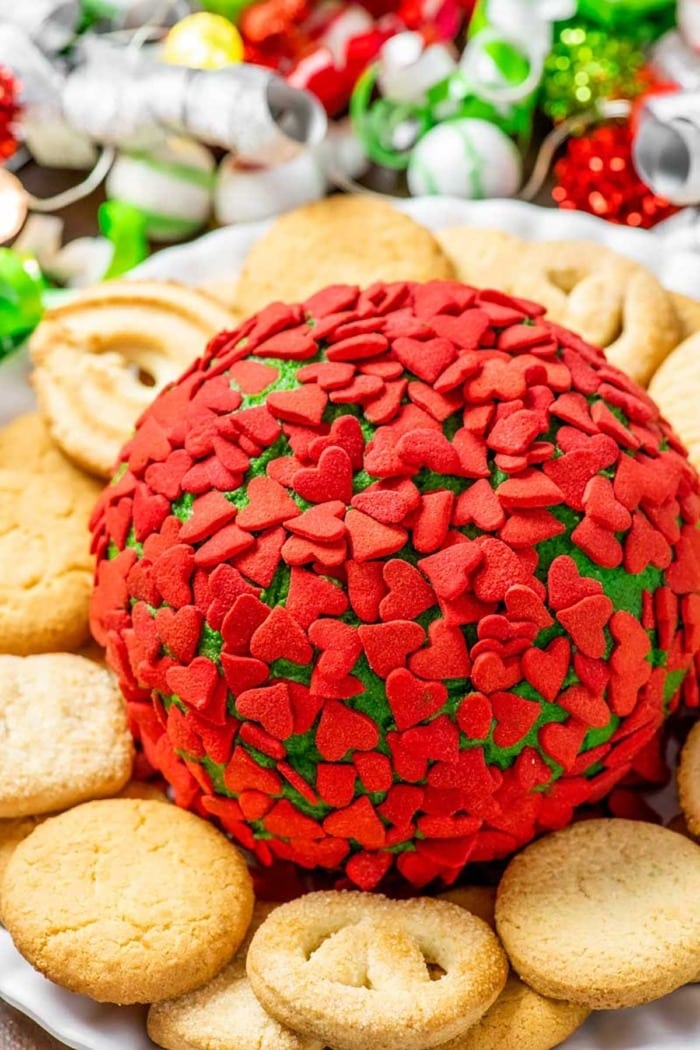 Grinch-themed cheese ball with green hue covered in tiny heart-shaped sprinkles on a cookie platter.