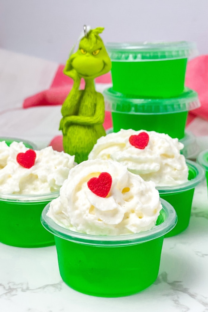 Green Jello shots on plastic cups with whipped cream and heart shaped candy garnish on top.