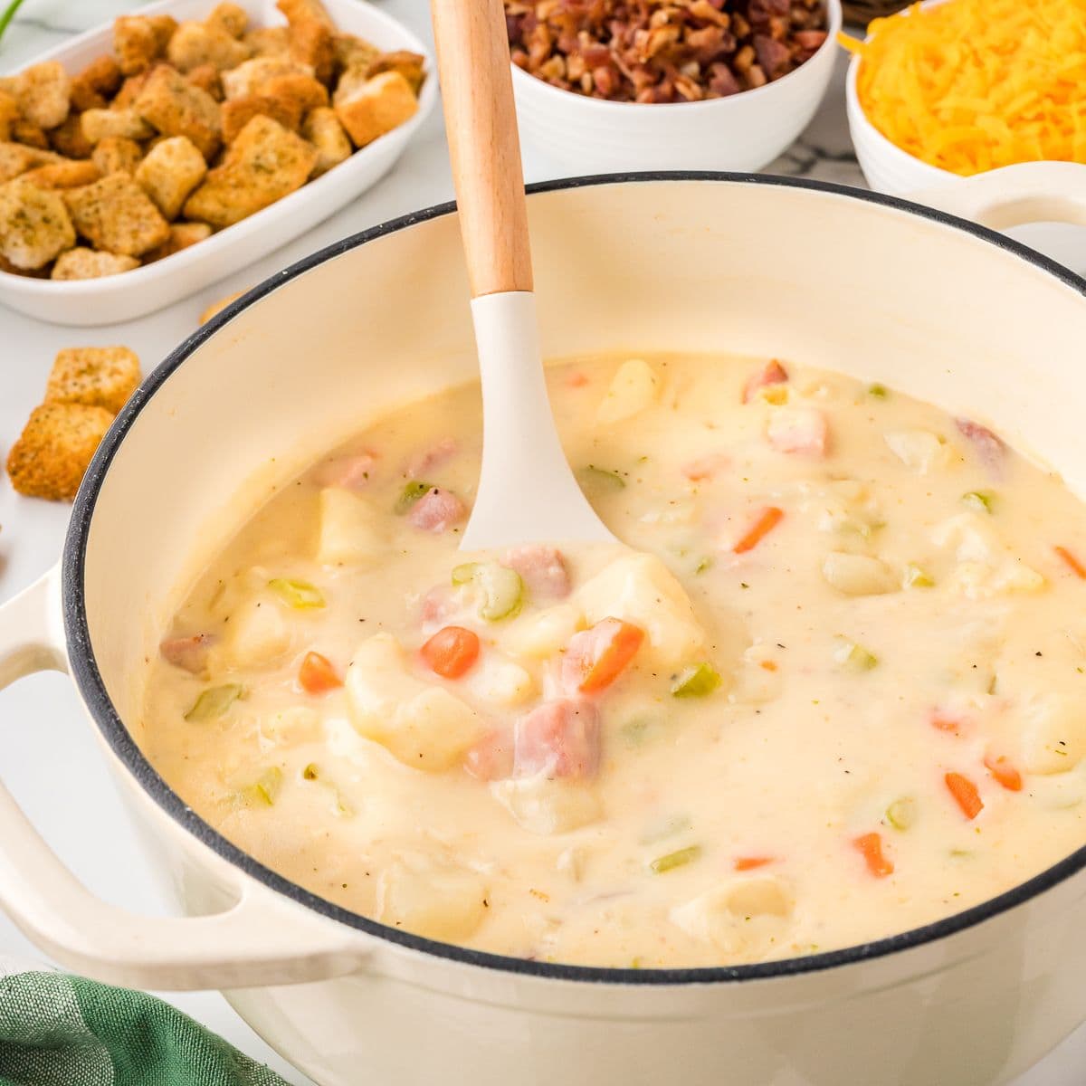 Creamy ham and potato soup in a pot with a wooden spoon and croutons, bacon bits, and shredded cheese on the side, close up