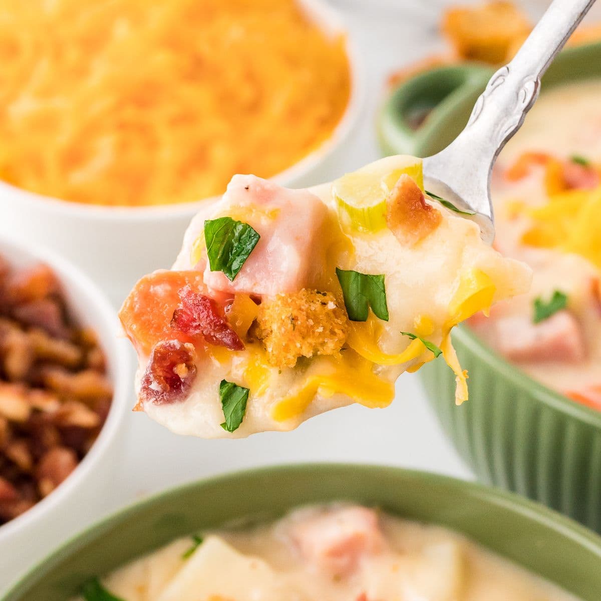 Spoonful of creamy ham and potato soup lifted from a bowl with chunks of ham, shredded cheese, and fresh herbs, close up on a spoon