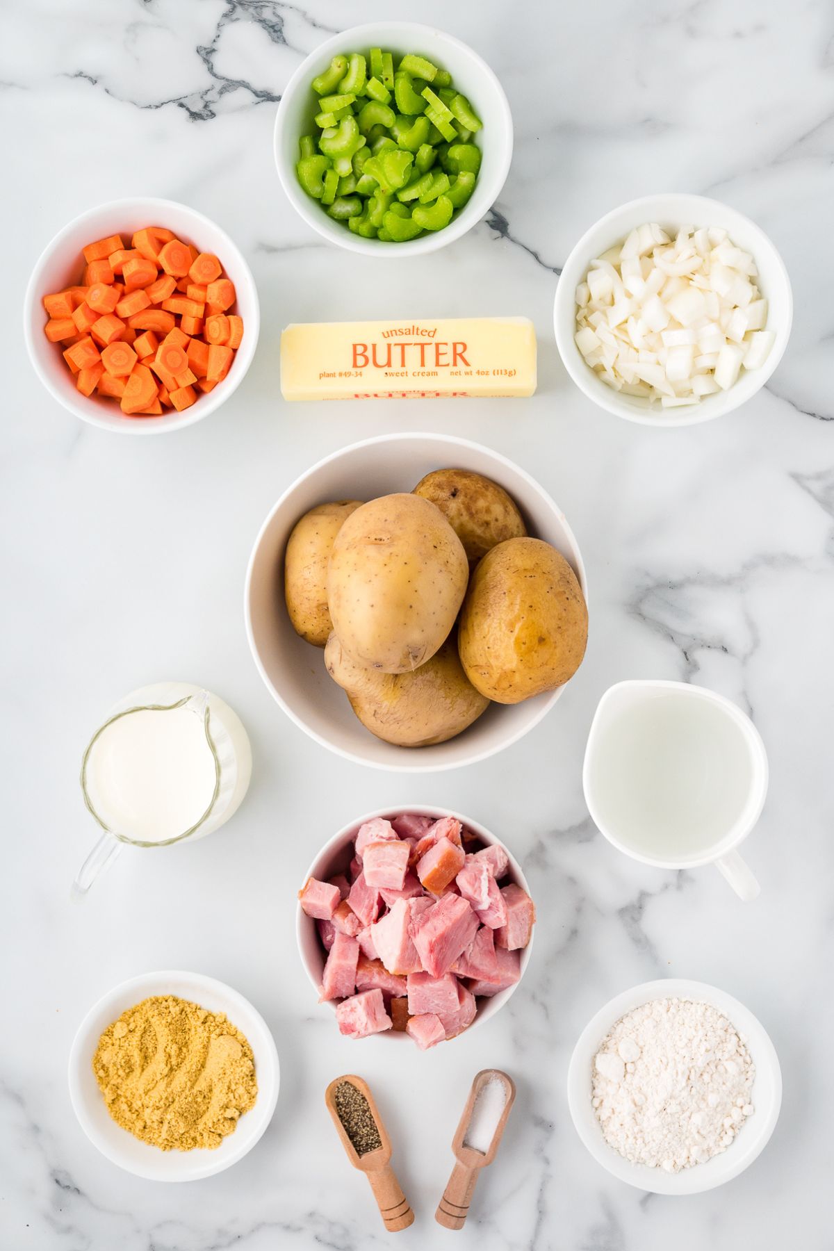 Ham and potato soup ingredients: Whole potatoes, sliced carrots, ham, diced celery, chopped onions, a stick of butter, milk, flour, salt, pepper, and chicken bouillon powder, top view.