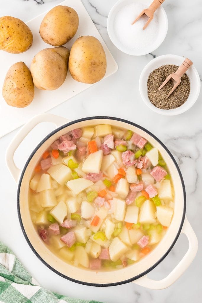 Diced potatoes, carrots, celery, onions, and ham in a Dutch oven with whole potatoes, salt, and pepper on the side, top view.