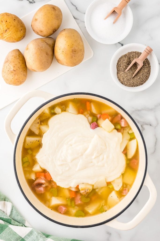 Ham, vegetable, and potato soup with creamy roux in a Dutch oven, with whole potatoes, salt, and pepper on the side, top view