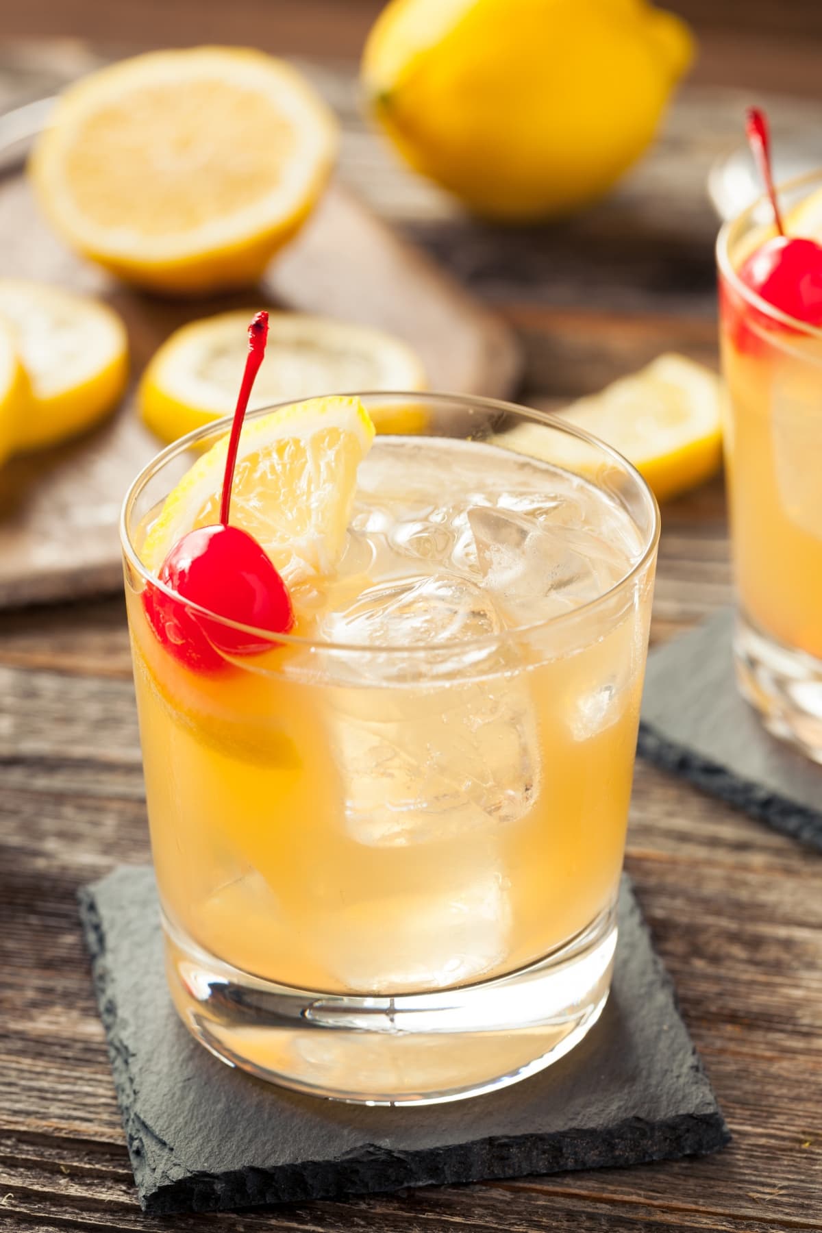15 Easy Tennessee Whiskey Cocktails featuring two glasses of homemade whiskey sour with ice, lemon, and cherry on slate coasters on a wooden table