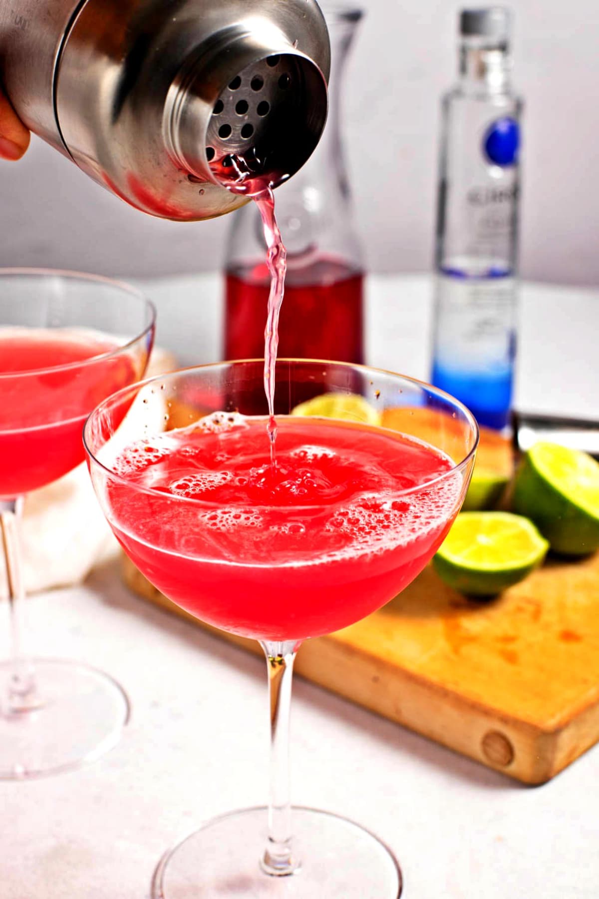 Cosmopolitan Cocktail Recipe (A Classic!) featuring A person pouring a pink cosmopolitan cocktail into a glass, with a full glass and a cutting board with limes behind it