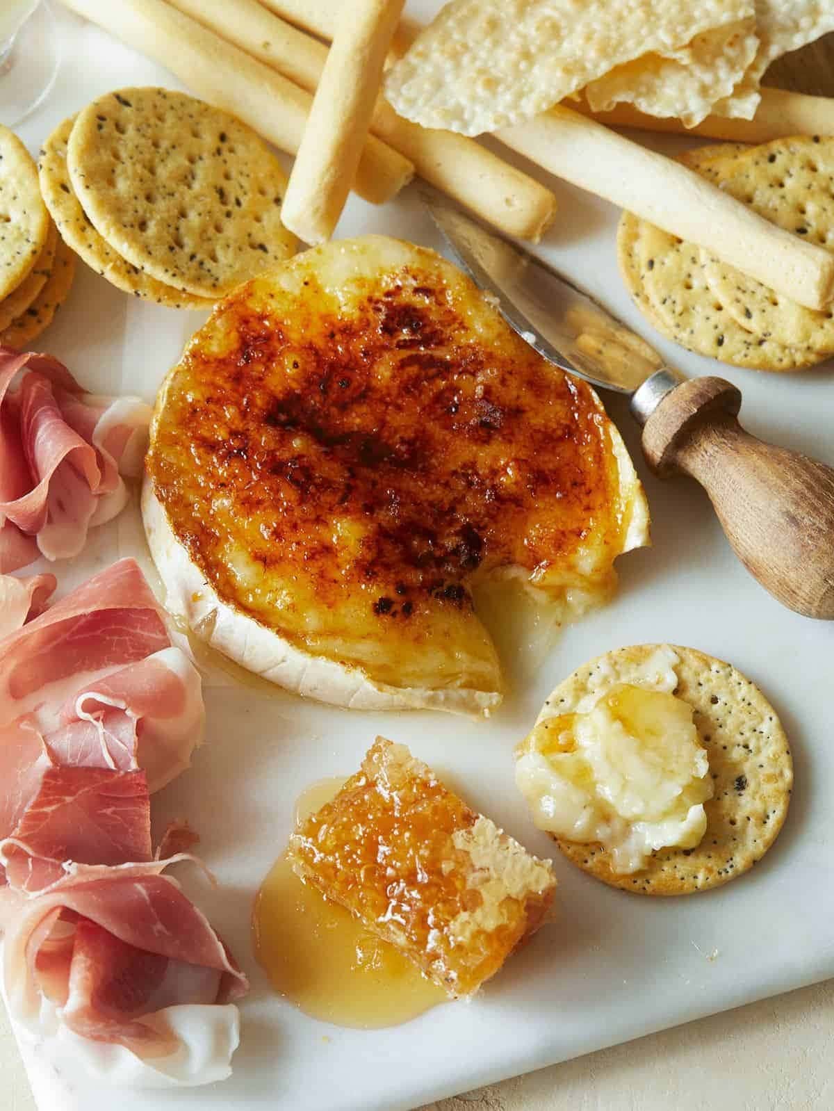 A cheese board with crackers, ham,cheese and honey brulee brie in the middle.