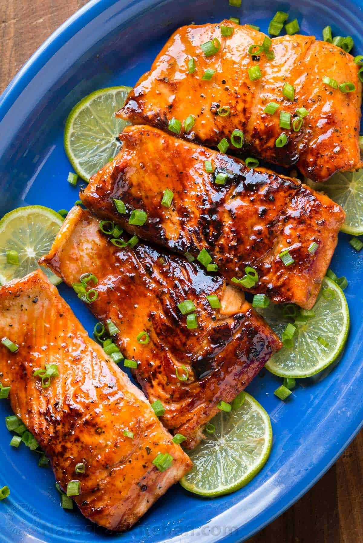 Honey glazed salmon belly garnished with chopped onion leaves and sliced lime served on a blue plate.